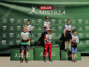 Powiększ obraz: Grupa zawodników na podium podczas dekoracji zwycięzców zawodów skoków narciarskich na tle baneru sponsorskiego