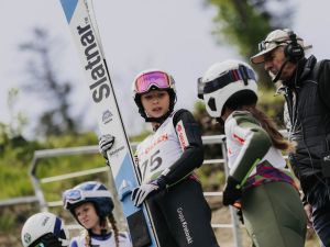 Powiększ obraz: Zawodnicy skoków narciarskich w kaskach i kombinezonach podczas rozmowy z trenerem przy skoczni