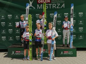 Powiększ obraz: Grupa młodych zawodników stojących na podium podczas ceremonii wręczenia nagród na tle zielonej ścianki sponsorskiej ORLEN